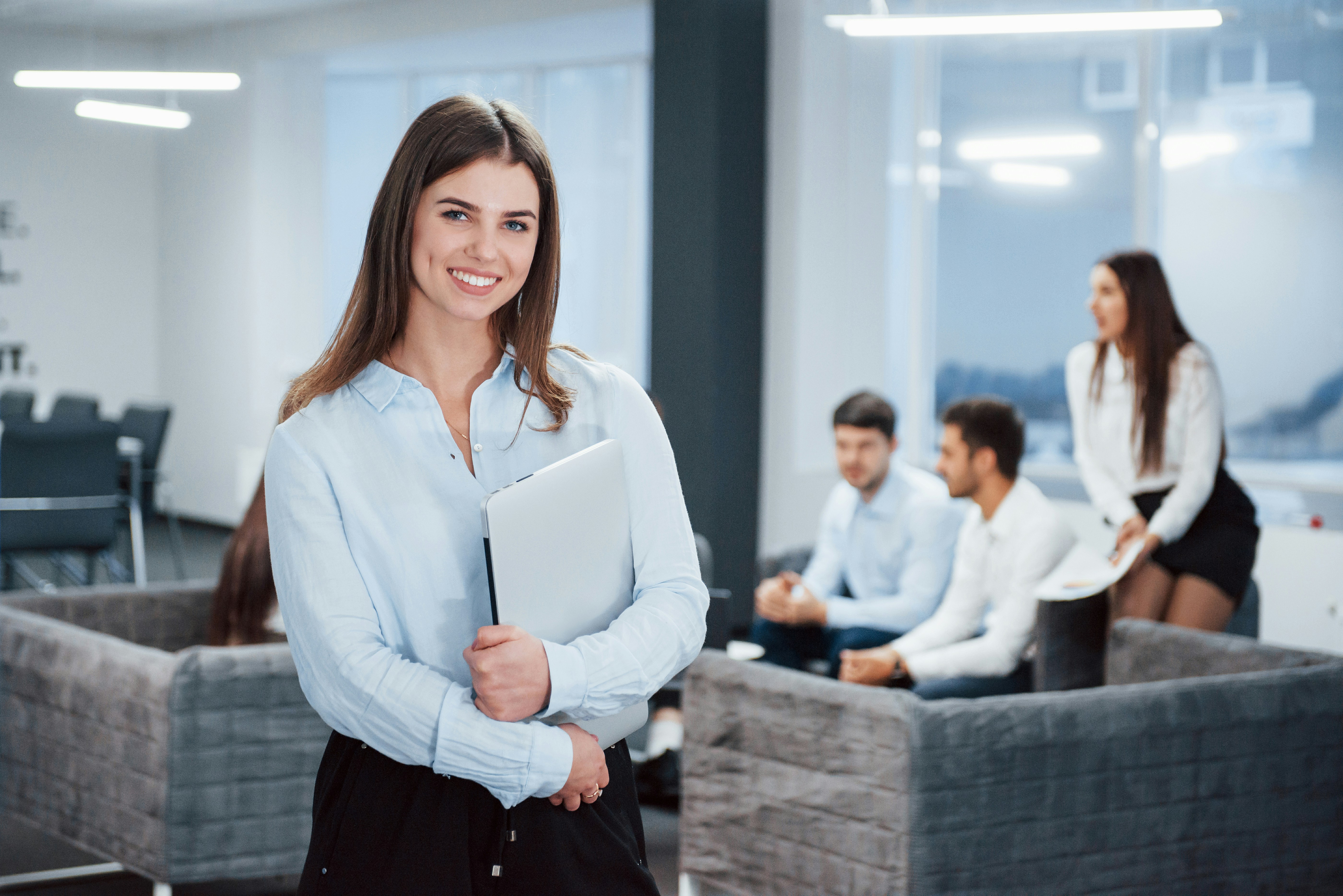  A temp worker standing in the office holding her laptop while a meeting is ongoing behind her.